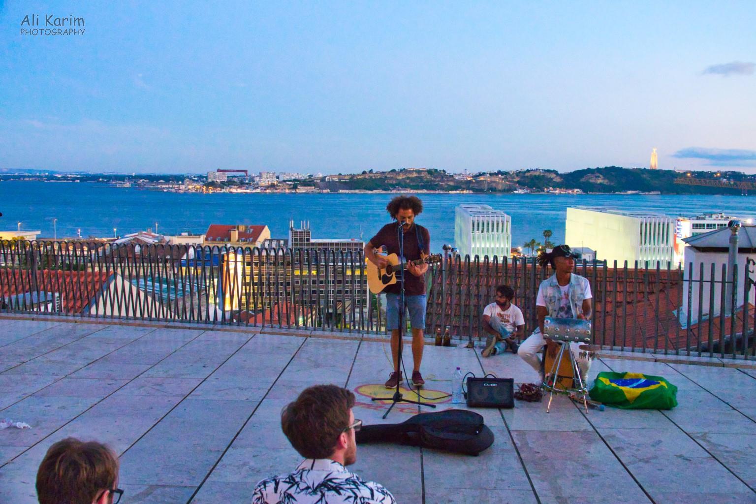 Lisbon Portugal: Entertainment at dusk at the Miradouro de Santa Catarina; one of multiple scenic viewpoints on the hills of Lisbon. You can see the Christo Rei statue in the background on the opposite riverbank, lit up for the evening.